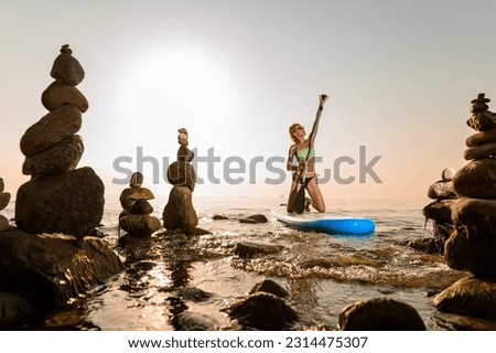 Beautiful girl sitting on paddle surf board at sea on stunning sunrise on background. Girl floating on sup surfing board between stones at sea