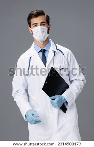 A male doctor in a white coat and a medical mask with a notepad in his hand looks at the camera on a gray isolated background, copy space, space for text