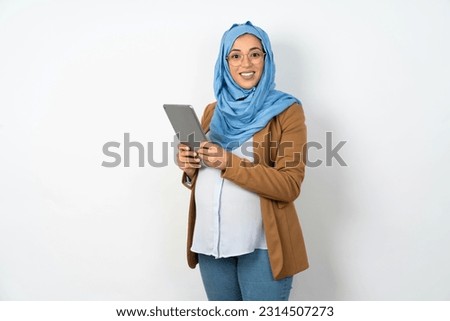 Photo of optimistic Young beautiful pregnant muslim woman wearing hijab over white studio background hold tablet. Teacher using modern technologies with school kids. 