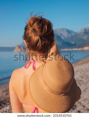 the girl stands with her back on her head, her hair is in a bun, she holds a hat in her hand, the mountains and the sea and the beach are in the background