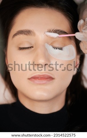 Woman with eye patch under lower eyelid keeping eyes closed while beautician applying cleansing mousse on eyebrows with lash brush. Female person having eyelash extension procedure in beauty salon. Royalty-Free Stock Photo #2314522575