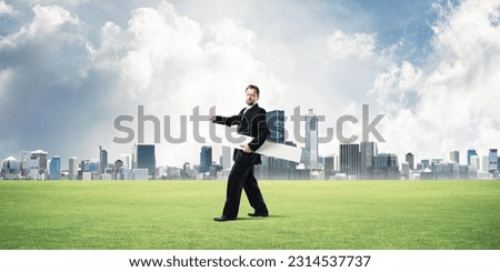 Confident and young businessman in suit holding big white arrow in hands which pointing to the side while standing on green lawn and cityscape view on background.