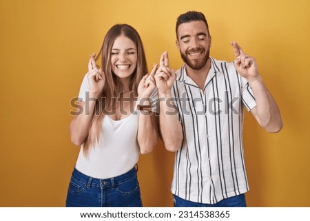 Young couple standing over yellow background gesturing finger crossed smiling with hope and eyes closed. luck and superstitious concept. 