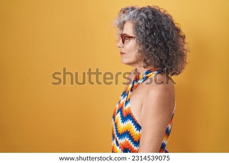 Middle age woman with grey hair standing over yellow background looking to side, relax profile pose with natural face and confident smile. 