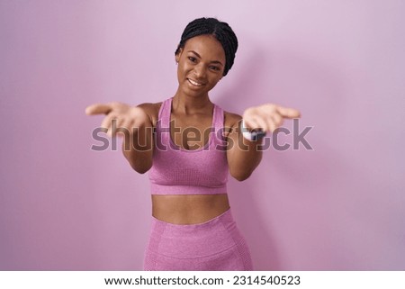 African american woman with braids wearing sportswear over pink background smiling cheerful offering hands giving assistance and acceptance. 