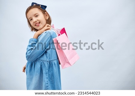 Pretty child girl in casual denim dress hold in hand pink shopping bags on white background, studio, Shopaholic shopping and fashion, discount sale concept, copy space, banner. Black friday