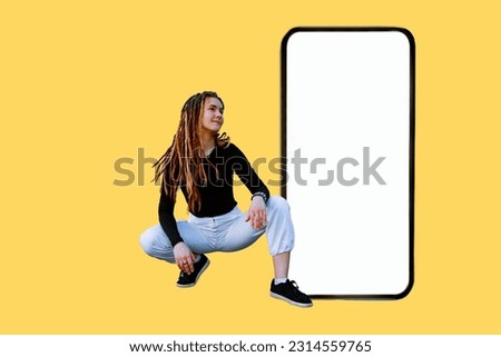 Demonstrating big smartphone screen, happy caucasian teen woman demonstrating big smartphone screen. Blank white display huge phone display mock up. Isolated on yellow, full body size sitting girl.