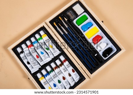 A landscape image of a rainbow paint set with brushes a pencil, eraser and sharpener in a wooden box on a beige background