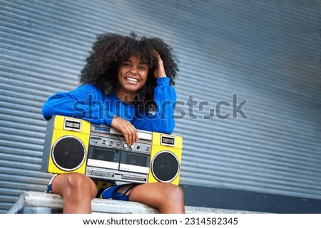 Portrait happy, cool young woman with curly hair holding yellow ghetto blaster at corrugated metal wall in alley Royalty-Free Stock Photo #2314582345