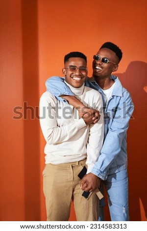 Portrait happy, stylish young gay male couple in sunglasses laughing and hugging on orange background
