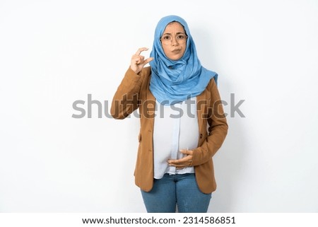 Displeased Young beautiful pregnant muslim woman wearing hijab over white studio background shapes little hand sign demonstrates something not very big. Body language concept.