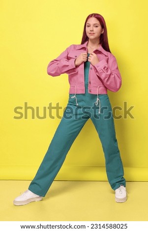A girl in a pink denim jacket and an oversized emerald jumpsuit on a yellow background studio isolated Royalty-Free Stock Photo #2314588025