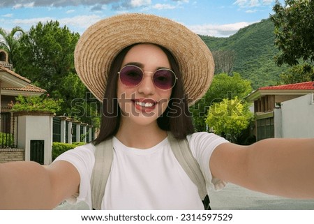 Smiling young woman in sunglasses and straw hat taking selfie in city