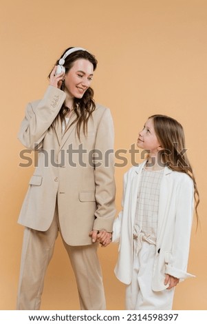 happy mother and daughter in suits, woman wearing wireless headphones near girl and holding hands on beige background, fashionable outfits, formal attire, corporate mom, modern family