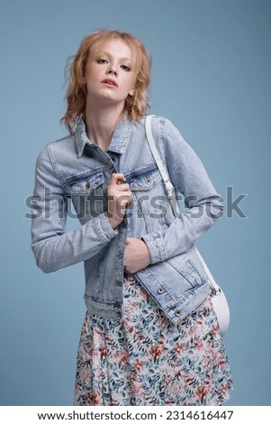 High fashion photo of a beautiful elegant young woman in a pretty colorful dress with floral pattern, denim, jeans jacket, handbag posing over blue background. Studio Shot, portrait. Blonde