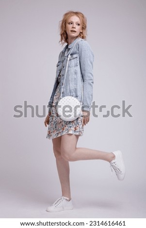 High fashion photo of beautiful elegant young woman in a pretty colorful dress with floral pattern, denim, jeans jacket, handbag posing over white, soft gray background. Studio Shot, portrait. Blonde