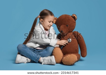 Little girl in medical uniform examining toy bear with stethoscope on light blue background