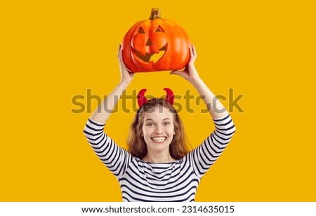 Happy woman holding up Halloween pumpkin. Cheerful beautiful young girl with devil horns standing isolated on yellow background, holding up carved pumpkin, looking at camera and smiling