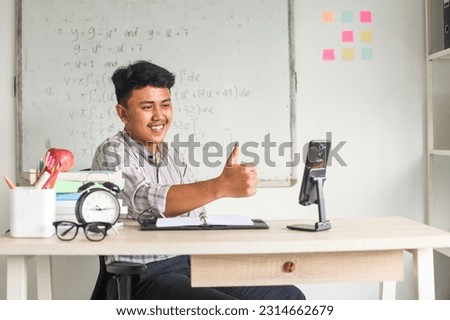 Education, online school and distant learning concept - Asian male math teacher having video call with students on smartphone screen and showing them thumbs up at classroom.