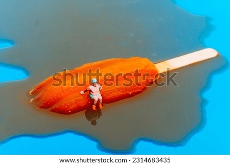 closeup of a miniature man wearing a green swimsuit, a green swimming cap and  swimming goggles sitting on a melting orange popsicle on a blue background Royalty-Free Stock Photo #2314683435