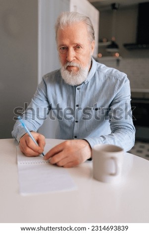 Vertical portrait of aged gray-haired man writing making notes in notebook, keep personal diary sitting at desk, looking at camera with serious expression. Mature male with pen and clipboard at home. Royalty-Free Stock Photo #2314693839