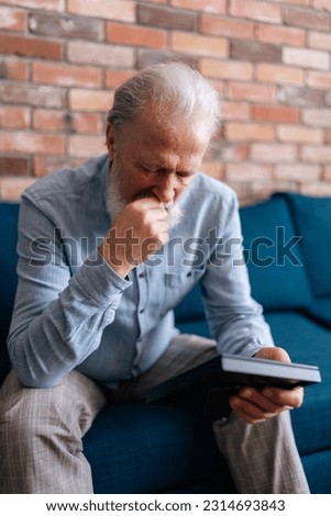 Vertical portrait of depressed elderly bearded man sitting on sofa at home, looking at picture of wife in frame and crying. Concept of nostalgia, grief, longing, loneliness in old age.