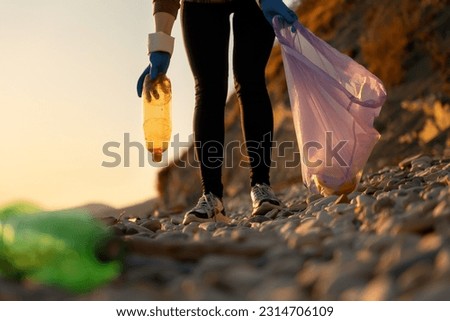 Cleaning of coastal areas from pollution. A volunteer walks along a pebble beach with a garbage bag and collects plastic bottles. Bottom view. The concept of preserving a sustainable ecosystem.