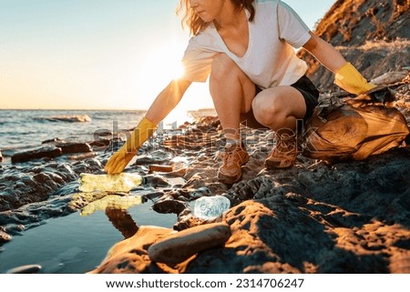 A young caucasian woman volunteer squats and picks up garbage on the ocean shore. Cleaning of the coastal zone. Concept of Earth Day. Royalty-Free Stock Photo #2314706247