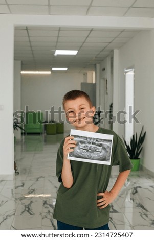 A smiling boy is standing in a dental clinic with a picture of his teeth in his hands. Portrait of a boy with an x-ray. Front view