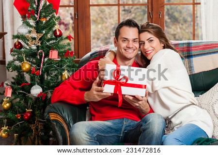 Celebrating Christmas together. Beautiful young couple sitting on the couch and smiling at camera exchanging gifts.