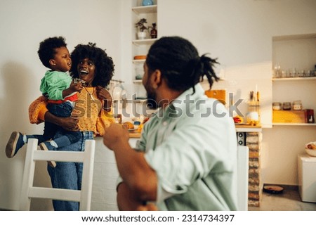 A multiracial mother is holding her beloved boy while standing in a kitchen at home while the husband is watching them and enjoying family time. A diverse family at home in the morning.