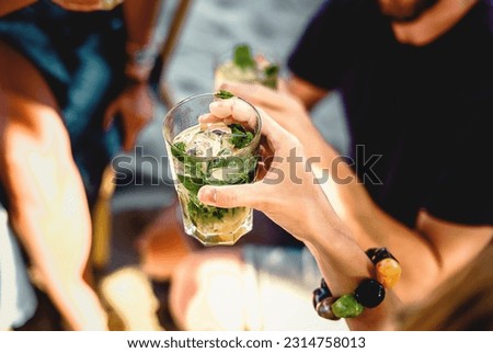 Refreshing Summer Mojito - Close-up of a hand holding a vibrant mojito cocktail with focus on ice. Friends enjoy the lively atmosphere in the background. Ideal for summer-themed projects.