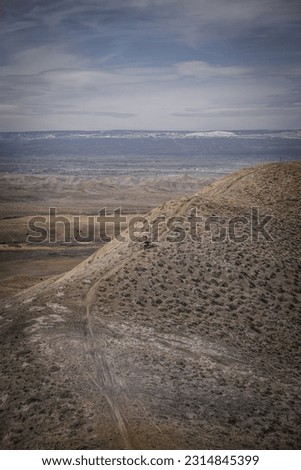 Far view of dirt bike rider on Trail on top of adobe clay ridgeline on hill in Peach Valley badlands in off road recreation area near Montrose Colorado with snow capped peaks in background 