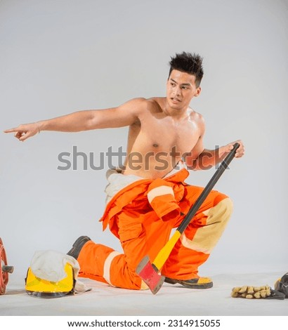Professional firefighter is sitting in a prepared position ready to save lives while firmly holding an iron axe and pointing finger to sideways.