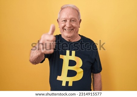 Senior man with grey hair wearing bitcoin t shirt approving doing positive gesture with hand, thumbs up smiling and happy for success. winner gesture. 