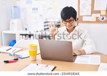 Young asian woman with short hair wearing call center agent headset doing ok sign with fingers, smiling friendly gesturing excellent symbol 