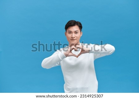 Portrait of attractive Asian man in red t-shirt speaks about own feelings, makes heart gesture over chest,