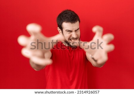 Young hispanic man wearing casual red t shirt shouting frustrated with rage, hands trying to strangle, yelling mad 