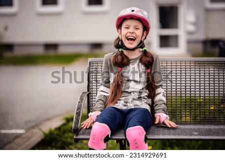 Schooler girl in helmet and full roller skating protection sittind on bench