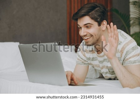 Young IT man wearing casual clothes t-shirt pajama lying in bed hold use work on laptop pc computer wave hand rest relax spend time in bedroom home in own room hotel wake up dream be in good mood day