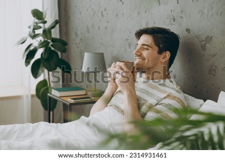 Profile happy young man wear casual clothes t-shirt pajama lying in bed drink coffee tea hold cup close eyes rest relax spend time in bedroom home in own room hotel wake up dream be in good mood day Royalty-Free Stock Photo #2314931461