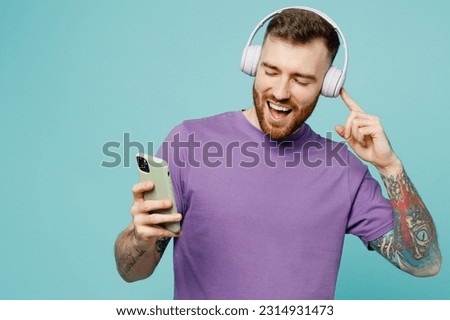 Young smiling happy man he wears purple t-shirt headphones listen to music use mobile cell phone isolated on plain pastel light blue cyan background studio portrait. Tattoo translates life is fight
