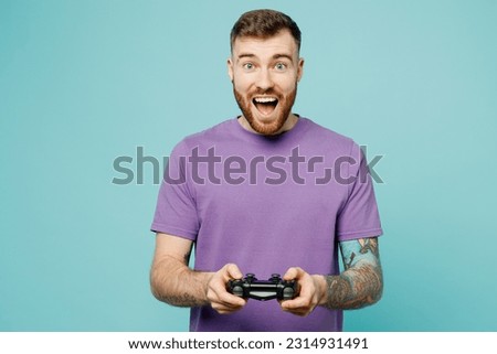 Young surprised excited man he wears purple t-shirt hold in hand play pc game with joystick console isolated on plain pastel light blue cyan background studio portrait. Tattoo translates life is fight