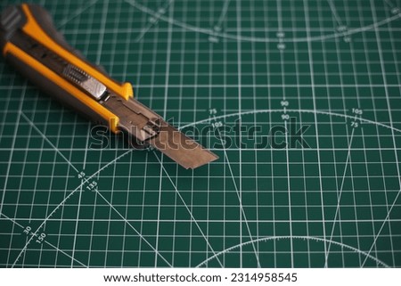 Stationery knife lies on a dark green cutting pad with marking lines for cutting paper and plexiglass.  Cutting board background with a sharp knife