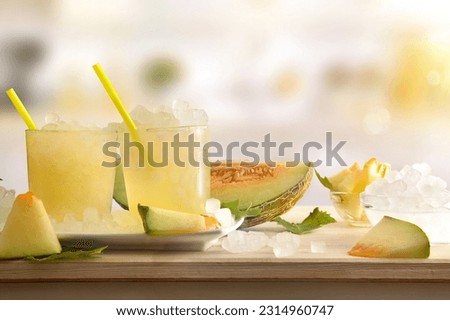 Two glasses with iced melon drink on wooden table full of crushed ice and sliced fruit around with kitchen in the background. Front view. Horizontal composition. Royalty-Free Stock Photo #2314960747