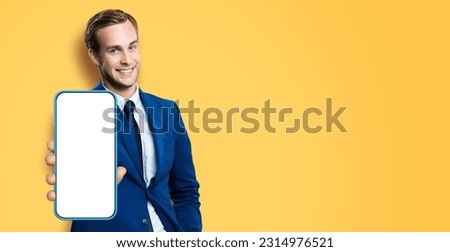 Wide image of smiling young business man, show cell phone, mobile smartphone. Happy businessman hold cellphone with white screen, isolated on yellow background. App advertisement concept.