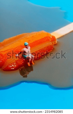 closeup of a miniature man, wearing a green swimsuit, a swimming cap and a pair of swimming goggles, sitting on a melting orange ice pop on a blue background Royalty-Free Stock Photo #2314982085