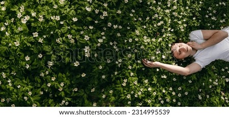 The portrait of beautiful cute woman no makeup in white shirt laying down on green grass with white flowers. Sunny summer day, wild nature, mood vacation, relax. Top view of girl. Be free, love forest Royalty-Free Stock Photo #2314995539