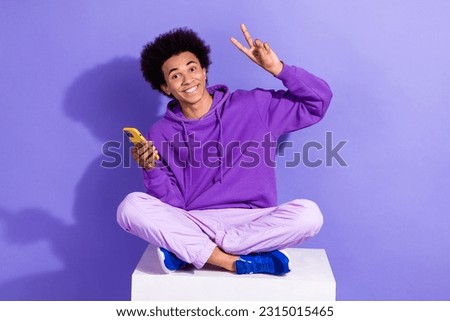 Full size photo of satisfied funky person violet hoodie sit on cube hold smartphone showing v-sign isolated on purple color background
