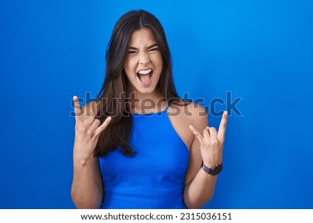 Hispanic woman standing over blue background shouting with crazy expression doing rock symbol with hands up. music star. heavy music concept. 